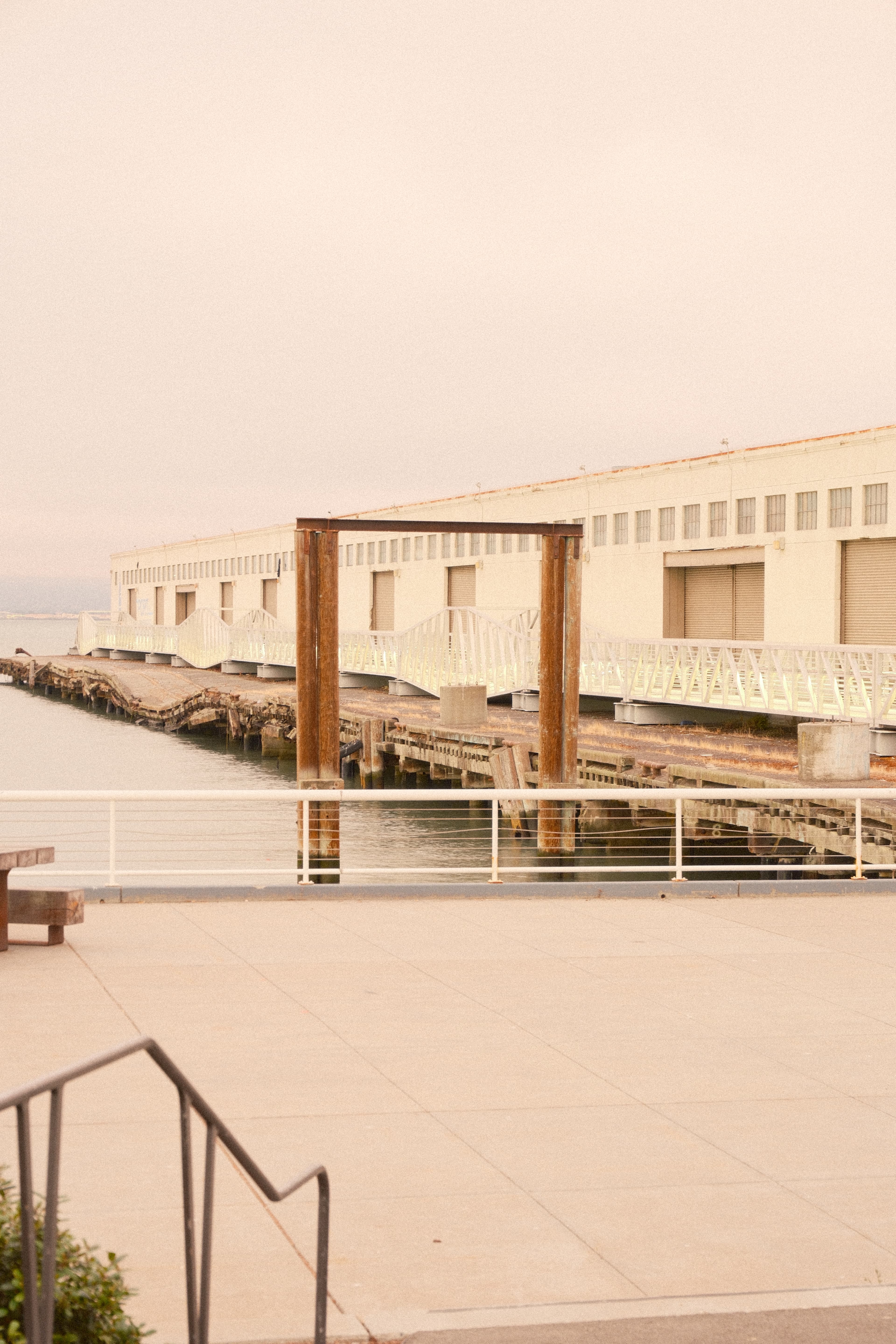 A long pier shed with white railings and weathered timber posts, in warm low light, with an empty concrete plaza in front.